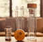 Two Azomalli-branded glass tea infusers, one fully assembled and one disassembled, sitting on a wooden table.