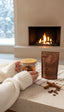 Cozy cacao tea moment in front of a fireplace. Cacao Spice pouch with tea mug brewing and cinnamon sticks and cacao beans nearby.