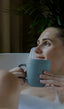 A light-skinned woman relaxing in a bathtub with a blue mug of tea.
